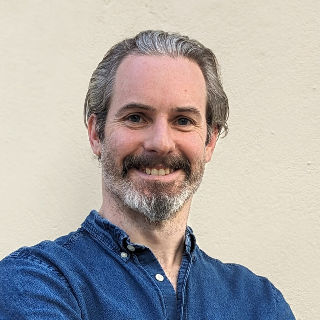 A portrait of Jamie Griffiths, a devilishly handsome man with greying hair and beard, in a blue shirt against a cream background.