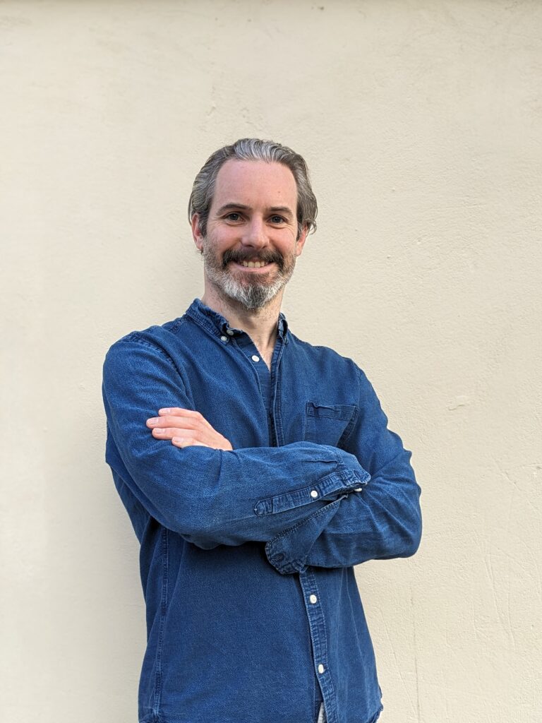 A smiling Jamie Griffiths, forty-three, in a blue shirt, leans against a beige wall, arms folded.